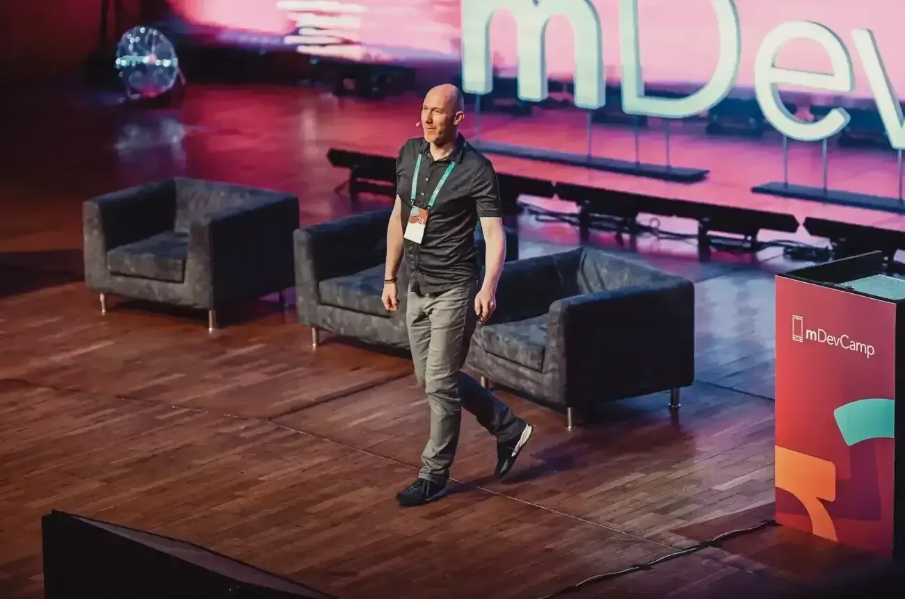 Marian Kamenistak walking the mDevCamp stage in front of a pink-lit conference backdrop.