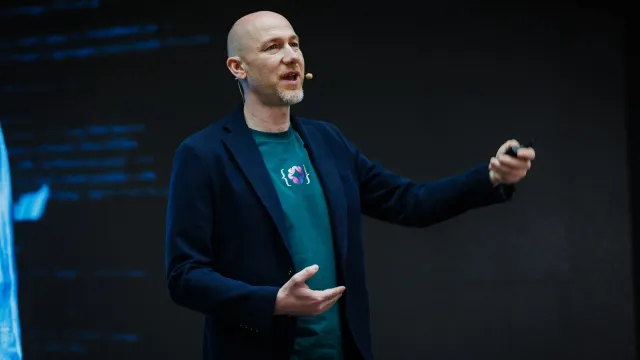 Marian Kamenistak mid-talk on a dark conference stage, lines of code projected on the screen behind him.
